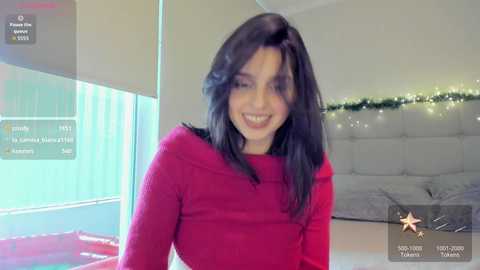 Video of a smiling woman with long black hair, wearing a red long-sleeve shirt, in a modern, festive room with a white tufted headboard, Christmas lights, and a red bedspread.