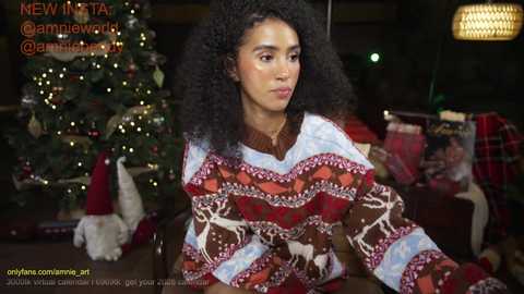 Video of a curly-haired woman in a festive sweater, sitting in a cozy, Christmas-decorated living room with a tree, presents, and a plaid blanket.