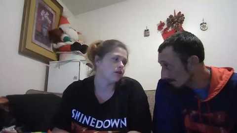 A video of two people in a casual indoor setting, the woman wearing a black \"SHINEDOWN\" shirt and the man in an orange hoodie, discussing in a cozy room with stuffed toys, framed art, and a white fridge.