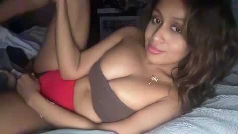 Video of a light-skinned woman with long brown hair, wearing a strapless brown top and red shorts, lying on a bed with a grey blanket.