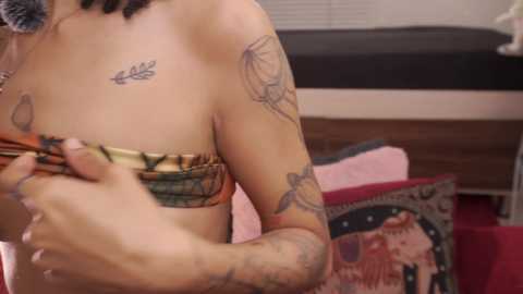 Video of a light-skinned woman with tattoos, including a dreamcatcher and a leaf, topless, adjusting a colorful bandeau in a bedroom with a bed, patterned pillows, and a tapestry backdrop.