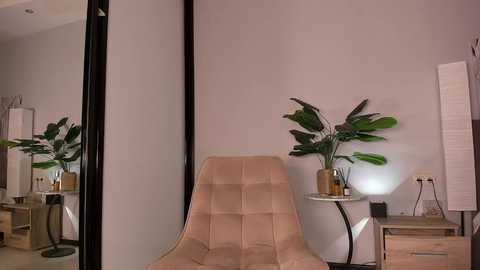 Video of a modern bedroom with a beige, tufted chaise lounge, a white side table, a tall floor lamp, and a potted plant; the room features neutral tones and a minimalist aesthetic.
