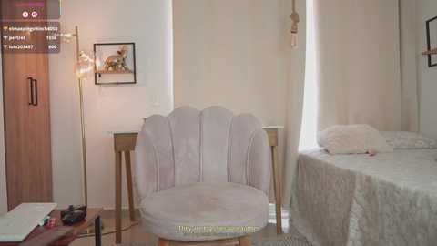 Video of a minimalist, beige-toned bedroom with a plush, cream-colored armchair, a small wooden nightstand, and a neatly made bed. A tall, floor lamp and a framed picture add subtle decor.