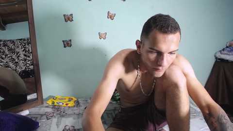 Video of a shirtless, light-skinned man with short brown hair, wearing maroon shorts, squatting on a bed with a leopard-print blanket, in a cluttered, pastel-colored room with butterfly stickers on the wall.