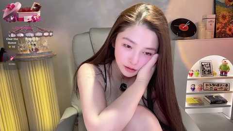 Video of a young Asian woman with long brown hair, wearing a black dress, sitting in a gray armchair, resting her head on her hand, looking thoughtful. The background features a bookshelf, a pink gift basket, and a record player.