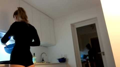 A video shows a woman with a ponytail, wearing a black long-sleeve shirt and orange shorts, washing dishes in a modern kitchen. The white cabinets, stainless steel sink, and bright lighting create a clean, minimalist ambiance.