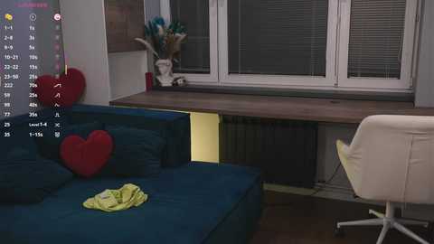 Video of a modern living room with a blue sofa, red heart pillow, yellow clothes on the floor, white office chair, wooden desk, and window blinds.