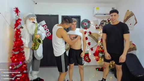 Video of a playful, festive indoor scene featuring two shirtless men and two men in black athletic wear, surrounded by Christmas decorations.