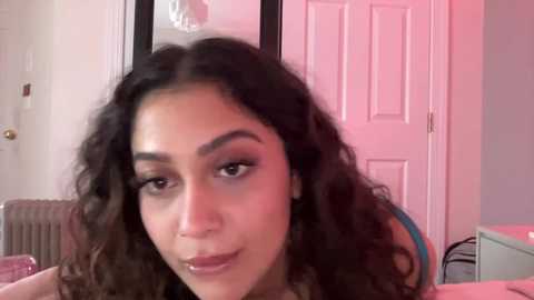 Video of a young woman with curly dark hair and light brown skin, smiling, in a pink-lit room with a closed white door and pink furniture.