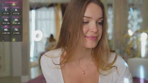 A video of a smiling, fair-skinned woman with long, straight, light brown hair, wearing a white top, and a delicate necklace, in a softly lit, elegant room with a large window.