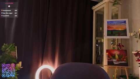 A video of a cozy room with a dark curtain backdrop, a lit ring light, a painting easel, and a colorful painting of a sunset landscape.