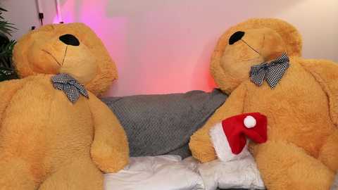Media: Video of two large, plush, light brown teddy bears with checkered bow ties, one in a red Santa hat, sitting on a gray and white quilted blanket against a soft pink wall.