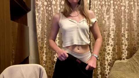Video of a light-skinned woman with blonde hair, wearing a white, off-the-shoulder crop top and black pants, pulling them down, in a room with beige curtains and a wooden chair.