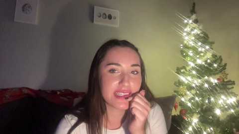 A video of a young woman with straight brown hair, smiling while holding a piece of broccoli, standing in front of a lit Christmas tree in a dimly lit room.