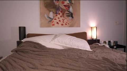 Video of a modern bedroom with a queen-sized bed, beige quilt, white pillows, and a large, colorful, abstract painting of a woman's face with red stars. Nightstands have modern lamps and personal items.