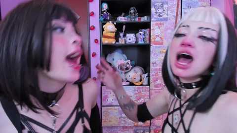 Video of two young women with pale skin and black hair, one in a black strappy outfit, the other in a black and white striped top, standing in a room with a pink and purple theme, filled with plush toys and anime decor.