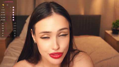 A video of a young Asian woman with long black hair and fair skin, wearing bold red lipstick, sitting on a bed in a dimly lit bedroom.