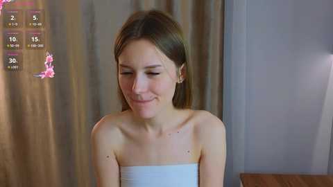 Video of a smiling young woman with fair skin, shoulder-length brown hair, and a light towel covering her shoulders, standing in a softly lit room with beige curtains.
