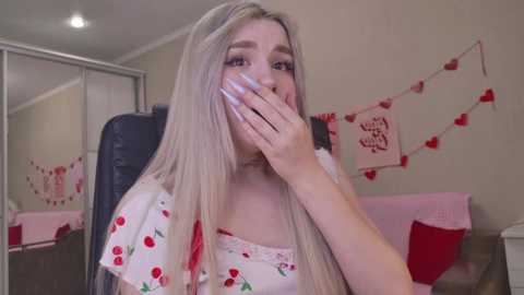 A video of a blonde woman with long hair, wearing a cherry-patterned dress, covering her mouth with her hand. The room has pink heart decorations and a white mirror.