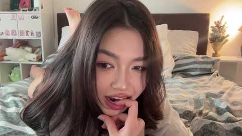 Video of an Asian woman with long black hair, lying on a bed with a striped blanket. She playfully touches her lips with a finger, looking seductively. Background shows a cluttered bedside table and a lit lamp.