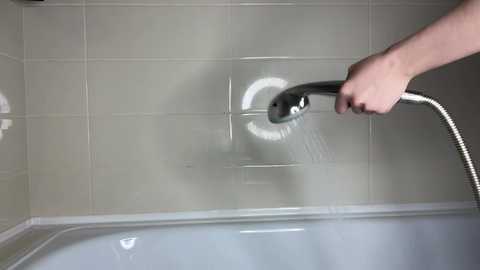 A video of a person's hand holding a handheld showerhead, spraying water onto a white bathtub with beige-tiled walls, creating a clean, minimalist bathroom scene.