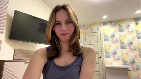 Video of a young Caucasian woman with straight, shoulder-length brown hair, wearing a blue tank top, standing in a brightly lit bathroom with pastel-colored, geometric-patterned wallpaper and a mounted flat-screen TV.