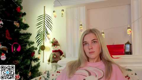 A video of a blonde woman in a cozy, festive bedroom with a lit Christmas tree, garland, and white bed.