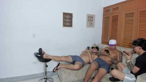 Video of three shirtless men in boxer shorts on a beige couch, watching a smartphone, in a minimalist room with white walls, wooden shutters, and a black stool.