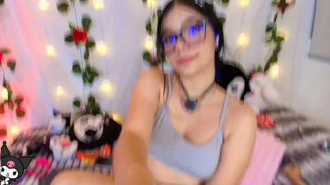 Video of a young woman with light skin, wearing glasses, a grey crop top, and a black choker, seated on a pink cushion. Background features white curtains adorned with fairy lights, pink and black pillows, and a panda toy.