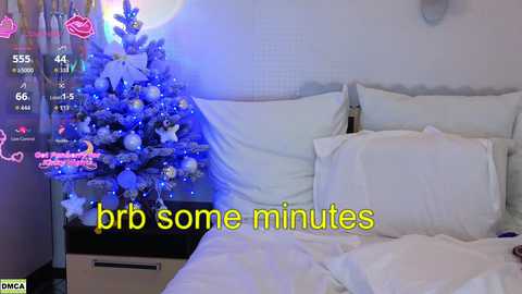 Video of a cozy bedroom with a white bed and a decorated Christmas tree in the corner, with a digital clock showing 4:44, and text overlaying \"brr some minutes\" in yellow.