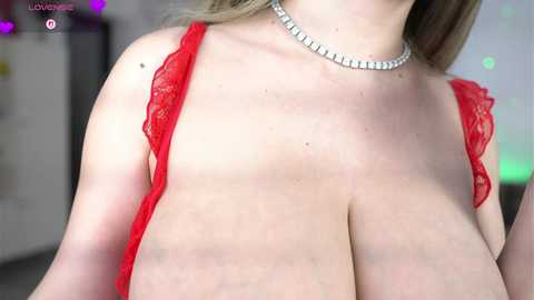 Video of a fair-skinned woman with large breasts, wearing a red lace bra and a silver choker necklace, standing indoors with blurred background.