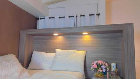 Video of a minimalist bedroom with a white bed, gray headboard, and a floral arrangement on a side table. Soft lighting, beige walls, and white curtains above the headboard create a cozy atmosphere.