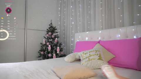 Video of a cozy bedroom with a white bed, pink and white pillows, a decorated Christmas tree, and twinkling fairy lights.