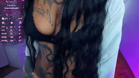 Video of a woman with long, wavy black hair, wearing a black bra, showcasing multiple tattoos on her chest and stomach. She is partially covered by a white shirt. Background features a purple-lit room with a calendar on the left.