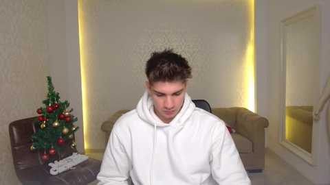 Video of a young man with short brown hair, wearing a white hoodie, standing in a modern, minimalist living room with a decorated Christmas tree and beige sofa.