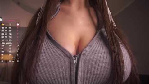 Media: Video of a woman with long, straight brown hair, wearing a grey ribbed sweater that reveals cleavage. Background shows a dimly lit room with a calendar and time display.