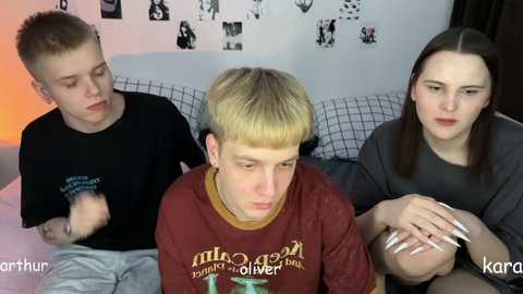 Video of three young people, two men and a woman, sitting on a bed, wearing casual clothes, smiling, with a white wall and black-and-white photos in the background.