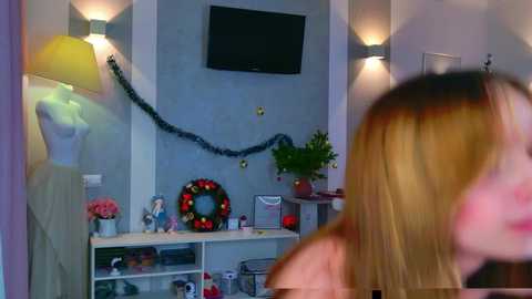 Video of a modern, well-lit room with a TV on a grey wall, a mannequin, festive decorations, a dollhouse, and a woman with blonde hair in the foreground.