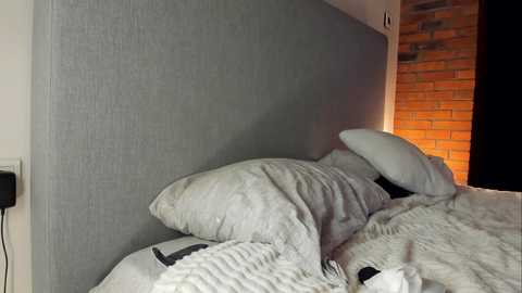 Video of a modern bedroom featuring a gray upholstered headboard, white duvet, and pillow, with exposed brick wall and soft lighting.