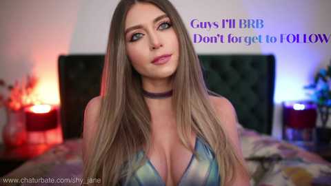 Video of a Caucasian woman with long, straight blonde hair, wearing a low-cut, blue and white patterned top, with a choker necklace. Background features a dimly-lit bedroom with a bed, candles, and a text overlay.