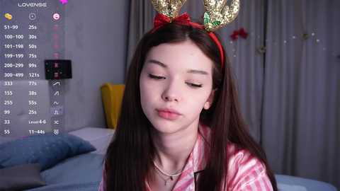 A video of a young Asian woman with fair skin, long brown hair, and closed eyes, wearing a red bow headband with golden rabbit ears, pink plaid shirt, and bed setting background.
