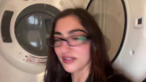 A video of a young woman with long dark hair, wearing glasses and a black top, standing in front of a washing machine. The background features a beige wall and a white door.