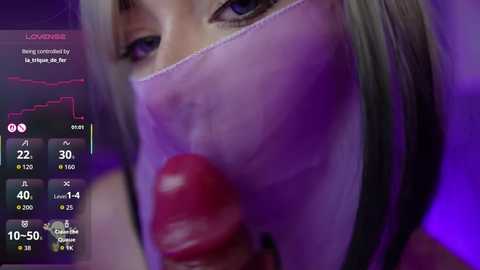 A video of a close-up of a woman with long blonde hair, wearing a face mask, holding a large, red, veiny dildo. The background features a graph with statistics, likely related to a virtual reality game.