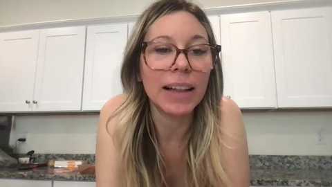 Video of a blonde, fair-skinned woman with glasses, wearing a light-colored top, leaning forward in a modern kitchen with white cabinets and a granite countertop.