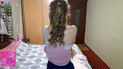 Video of a woman with long, wavy brown hair in a white tank top and dark pants, sitting on a bed with a grey polka-dot comforter in a simple, beige-walled room.