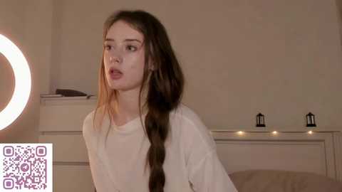 Video of a young girl with long, straight brown hair, wearing a white t-shirt, looking surprised. Background includes a white dresser with two black lanterns and a ring light.