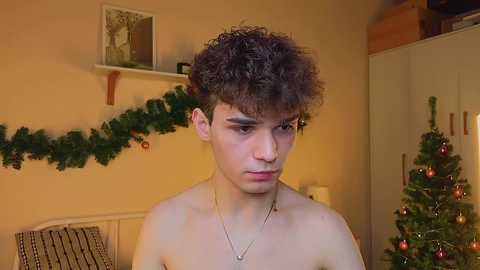 Video of a young, shirtless, curly-haired, light-skinned boy in a cozy room with a decorated Christmas tree and garland.