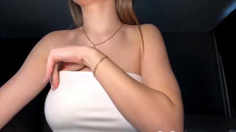 Media: Video of a woman with light skin, long straight brown hair, wearing a strapless white dress, adjusting her dress with one hand, in a dimly lit room with dark walls.