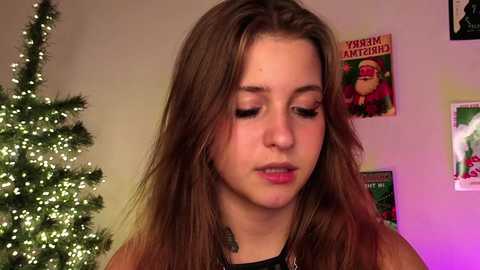 Video of a young, fair-skinned woman with long, brown hair, wearing a black top, standing next to a decorated Christmas tree with twinkling lights. In the background, festive posters and a red and green Santa Claus ornament.
