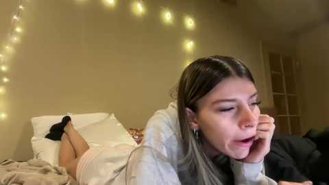 Video of a young woman with long brown hair, wearing a gray hoodie, lying on a bed with white pillows and beige sheets. She's resting her head on her hand, looking relaxed in a dimly lit room with fairy lights.